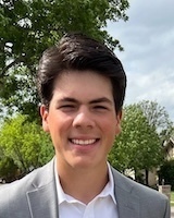 Headshot of a young man with dark hair, wearing a gray suit and white shirt, smiling outdoors with trees in the background.