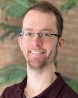 A man with glasses and a beard smiles, wearing a dark collared shirt. Blurred brick wall and green foliage are in the background.