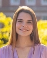 A young woman with long brown hair smiles, wearing a light purple top outdoors.