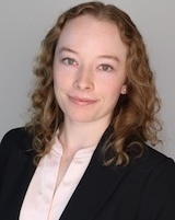 A woman with curly brown hair and a slight smile wears a black blazer and a light pink blouse.
