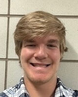 A young man with light brown hair and a wide smile wears a blue and white plaid shirt.
