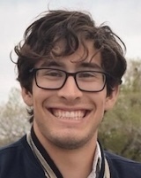 A young man with dark, curly hair and black-rimmed glasses smiles broadly, showing teeth. He wears a navy blue jacket.