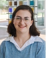 Smiling woman with dark hair and light-rimmed glasses wears a blue v-neck sweater over a white collared shirt. Behind her, a structure features colorful rectangular glass panels.