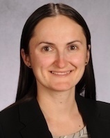 A woman with long dark hair and a slight smile wears a dark blazer over a light patterned top, against a muted background.