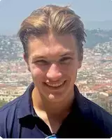 Young man with blonde hair and a dark blue polo shirt smiles, with a sunny, hilly coastal town in the background.