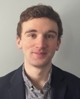 A young man with brown hair and light eyes smiles subtly, wearing a dark navy blazer over a light patterned shirt.