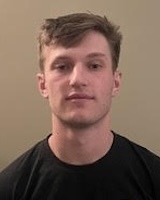 Headshot of a young man with short brown hair, light skin, and a neutral expression, wearing a dark t-shirt against a plain background.