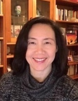 An East Asian woman with dark, shoulder-length hair smiles, wearing a dark grey turtleneck. Behind her are wooden bookshelves packed with books.